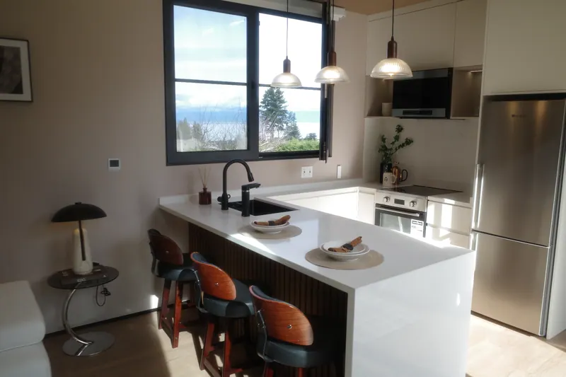 Light-finish kitchen with a quartz waterfall peninsula, three stools and an ocean view through the window