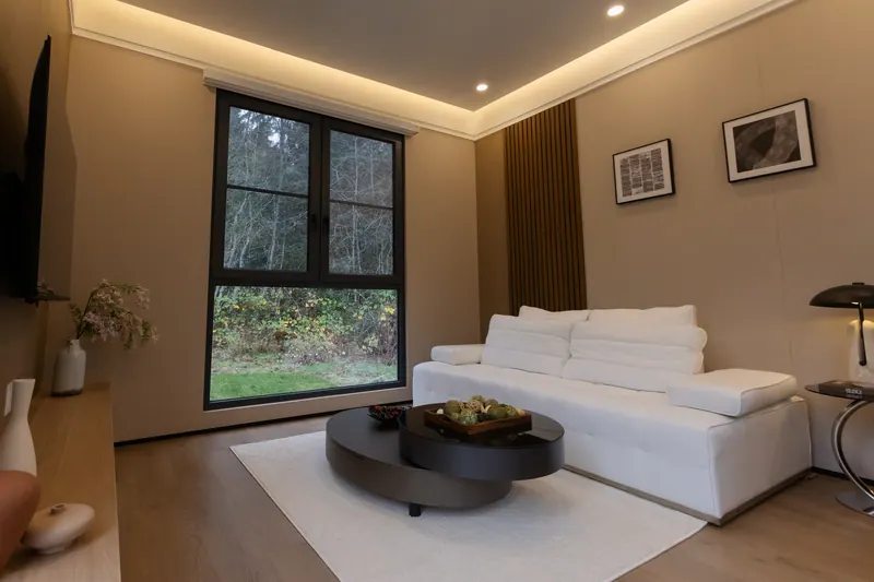 Light-finish living room with a white sofa, cove lighting and a tall window onto the forest