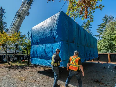 Two crew members beside a Cueva home under a blue weather tarp as the crane sets it in place